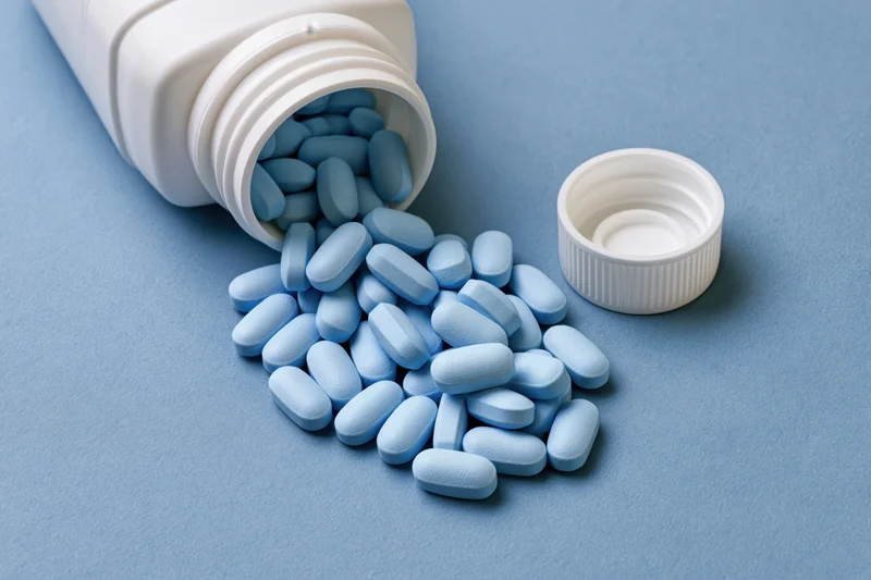 Light-blue oval pills spilling from a white plastic bottle onto a blue surface