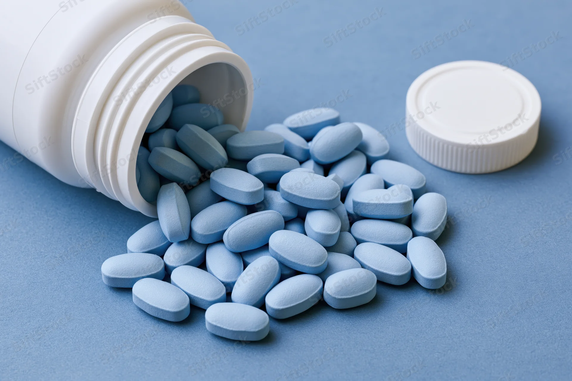 Light-blue oval pills spilling from a tipped white bottle onto a blue surface