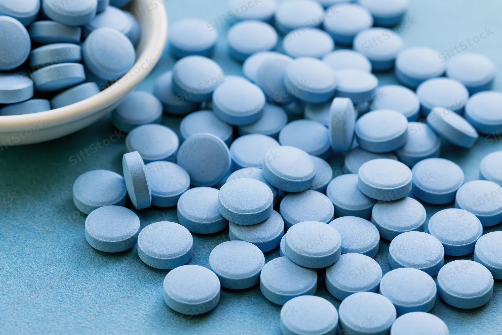 Light-blue pills scattered across a flat surface with a ceramic bowl in the corner