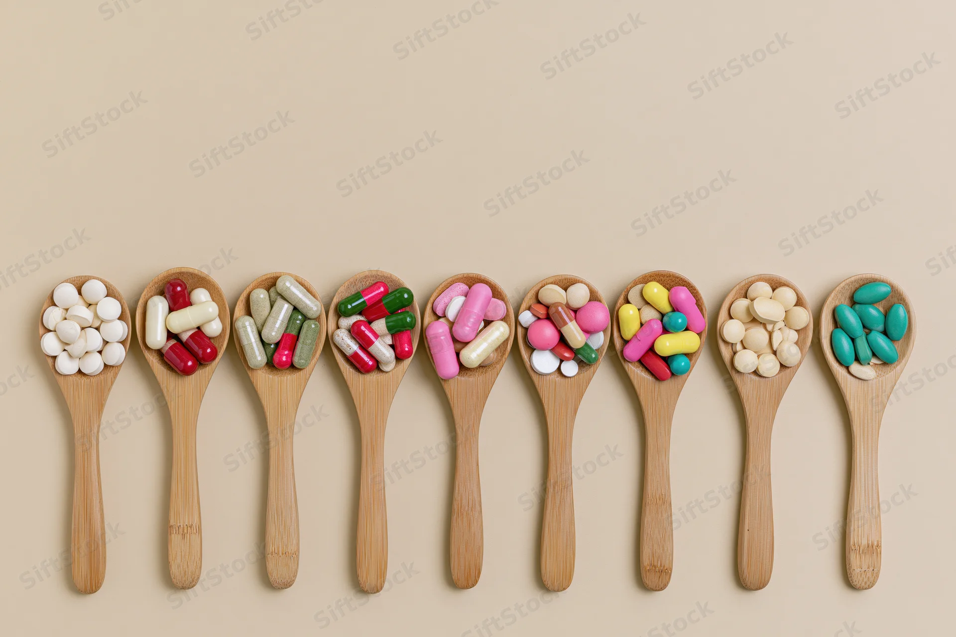 Eight wooden spoons each holding different colored pills arranged in a row on a beige background
