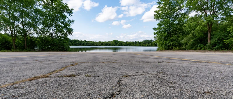 Empty asphalt parking lot facing calm lake surrounded by trees under blue sky with clouds