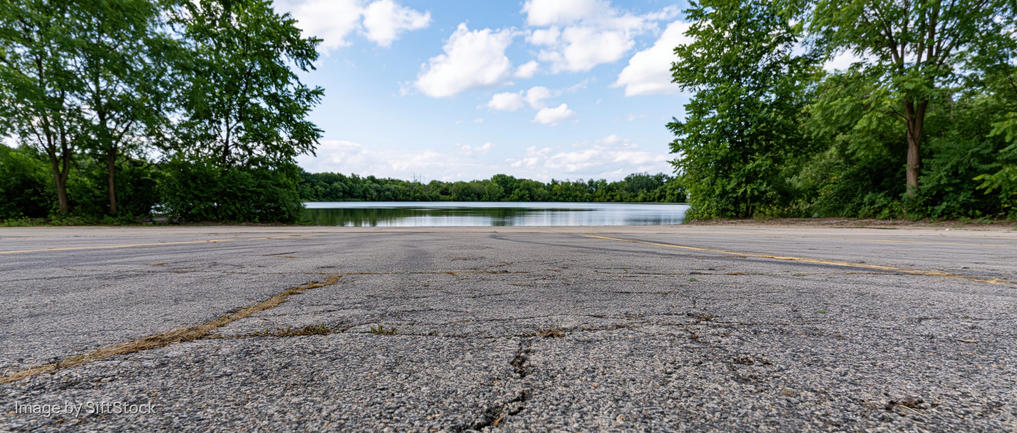 Empty asphalt parking lot facing calm lake surrounded by trees under blue sky with clouds