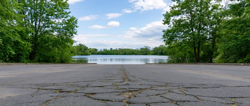 Empty asphalt parking lot facing calm lake surrounded by green trees under blue sky with clouds