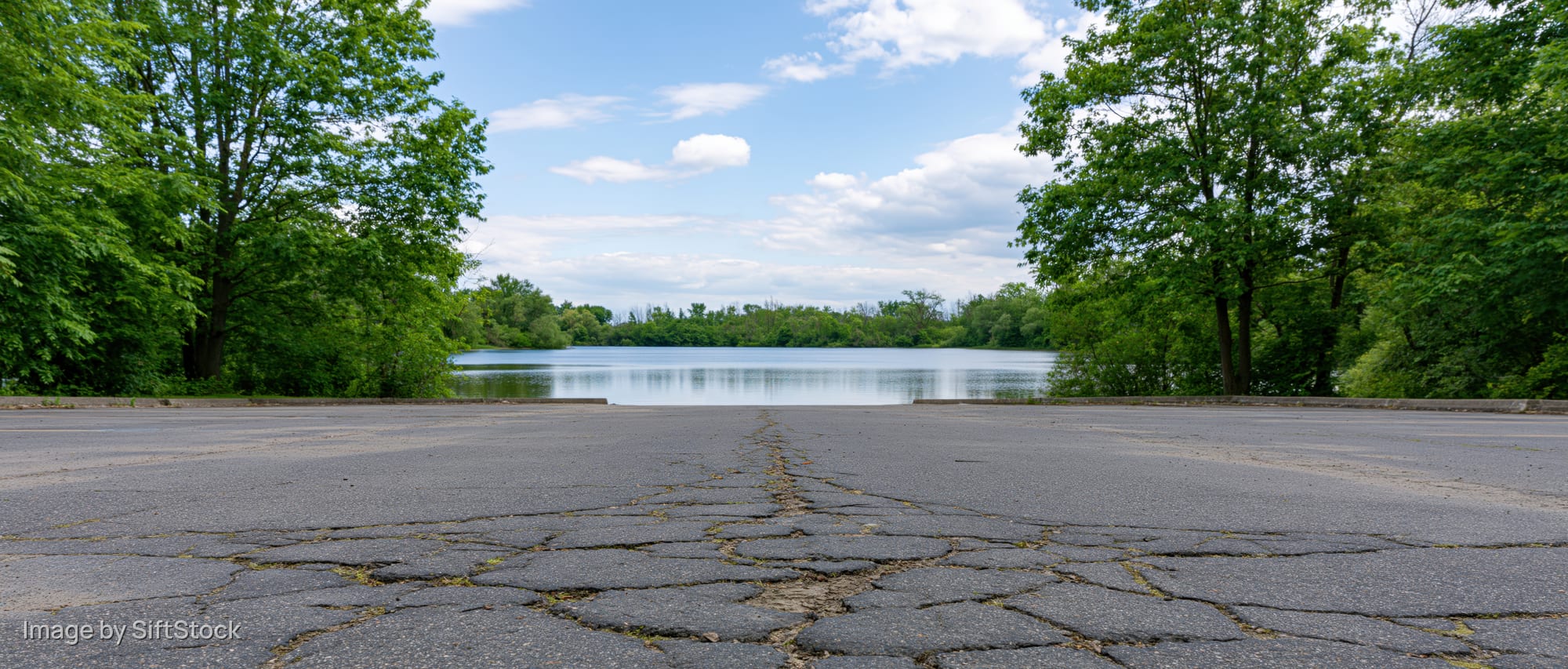 Empty asphalt parking lot facing calm lake surrounded by green trees under blue sky with clouds