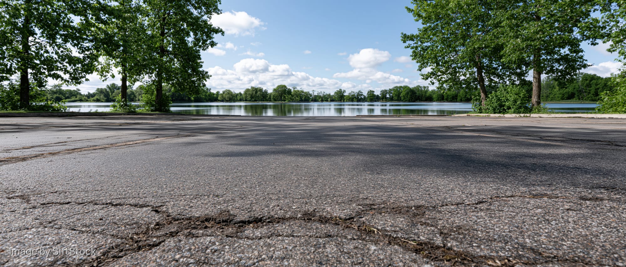 Empty asphalt parking lot facing calm lake with trees and sky in background