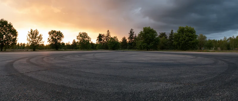 Curved tire skid marks on empty asphalt racing circuit at sunset with trees and cloudy sky in background