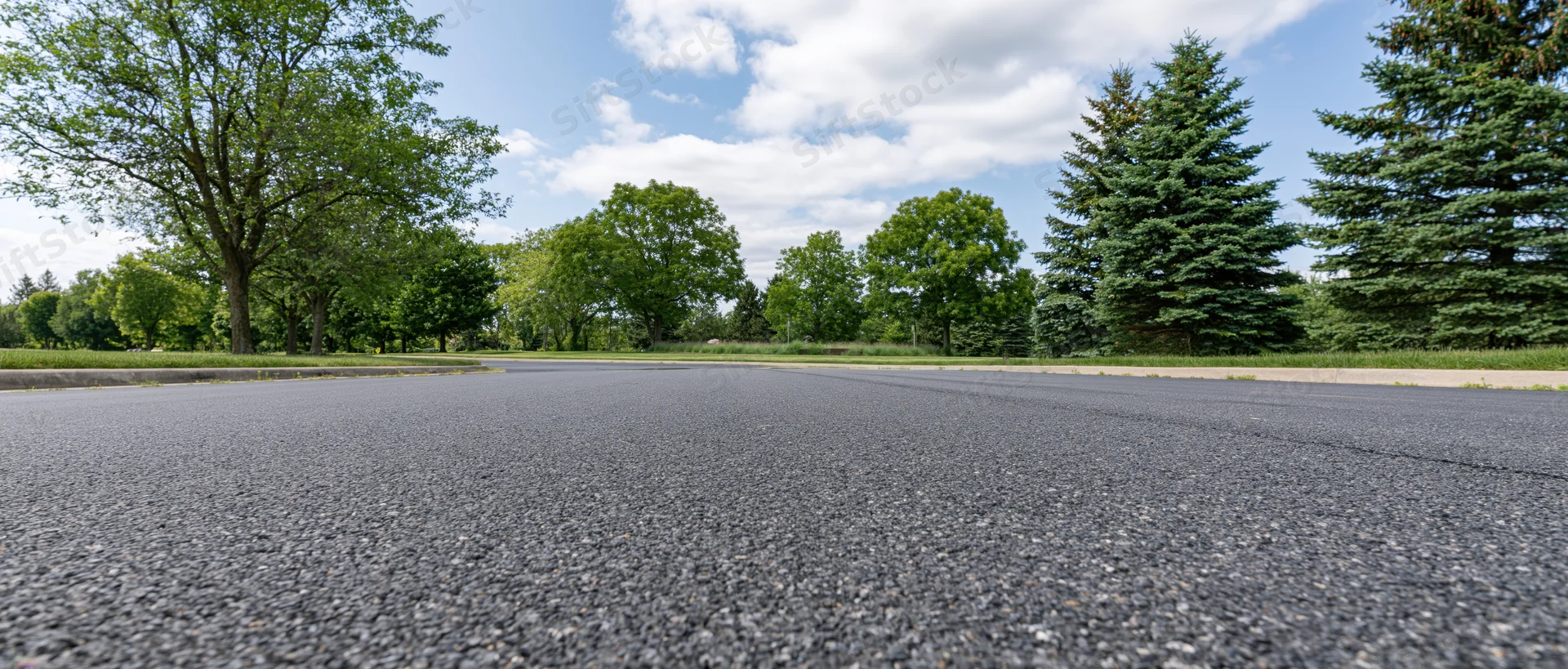 Low-angle view of empty asphalt road stretching toward tree-lined horizon under partly cloudy sky