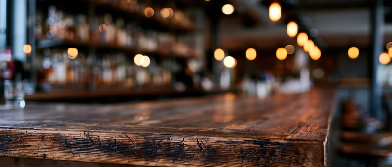 Dark wooden bar counter in sharp focus with blurred bottles and warm lights behind