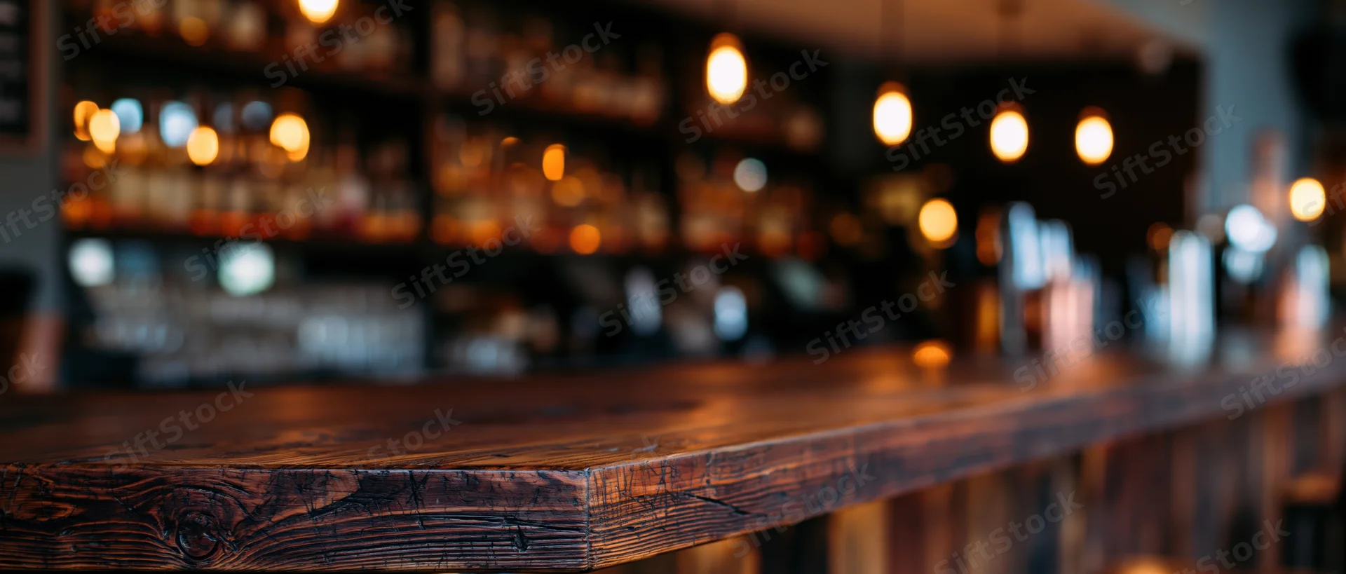 Dark wood bar counter in sharp focus with blurred background of bottles and pendant lights