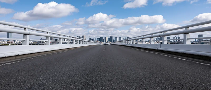 Empty elevated highway overpass with white concrete guardrail leading toward city skyline under blue sky with clouds