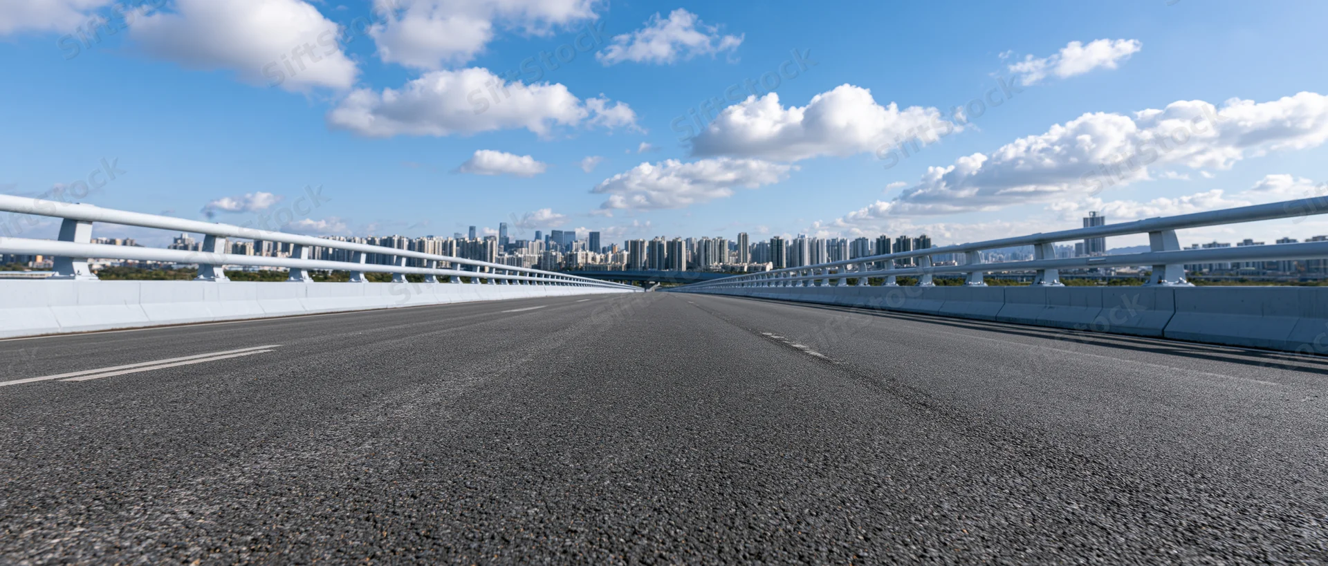Empty elevated highway overpass with white concrete guardrail leading toward city skyline under blue sky with clouds