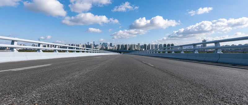 Empty elevated highway overpass with white concrete guardrail leading toward city skyline under blue sky with clouds