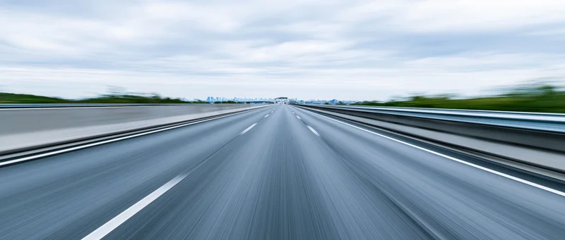 Drivers-eye view of empty elevated highway stretching toward distant city skyline under overcast sky