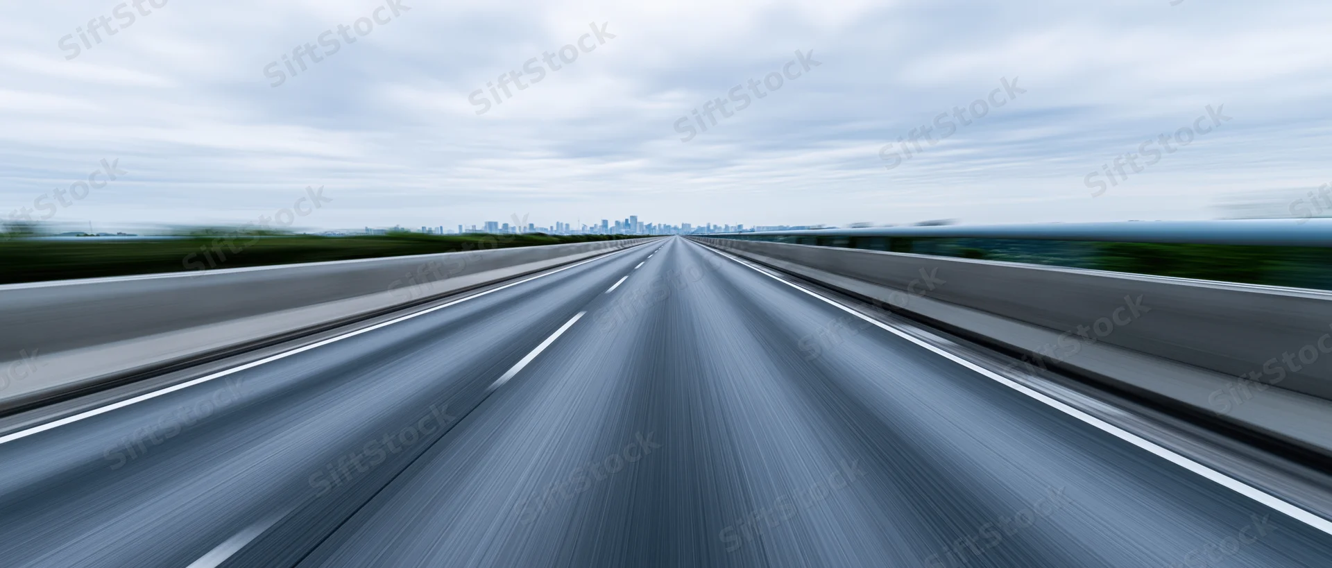 Drivers-eye view of an elevated highway stretching toward a distant city skyline under an overcast sky