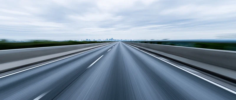 Drivers-eye view of an elevated highway stretching toward a distant city skyline under an overcast sky