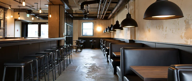 Empty industrial-style restaurant interior with booths, bar stools, and pendant lights along a concrete floor