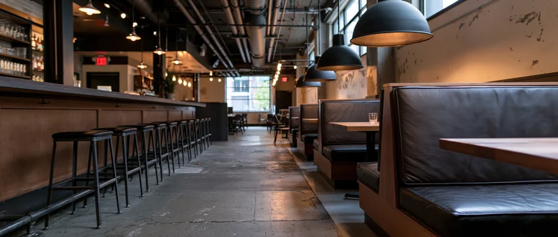 Empty industrial-style restaurant interior with booths and bar stools along concrete floor and exposed ceiling pipes