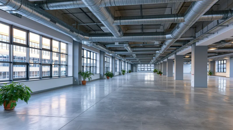 Empty industrial interior with exposed metal ductwork and large windows along concrete floor