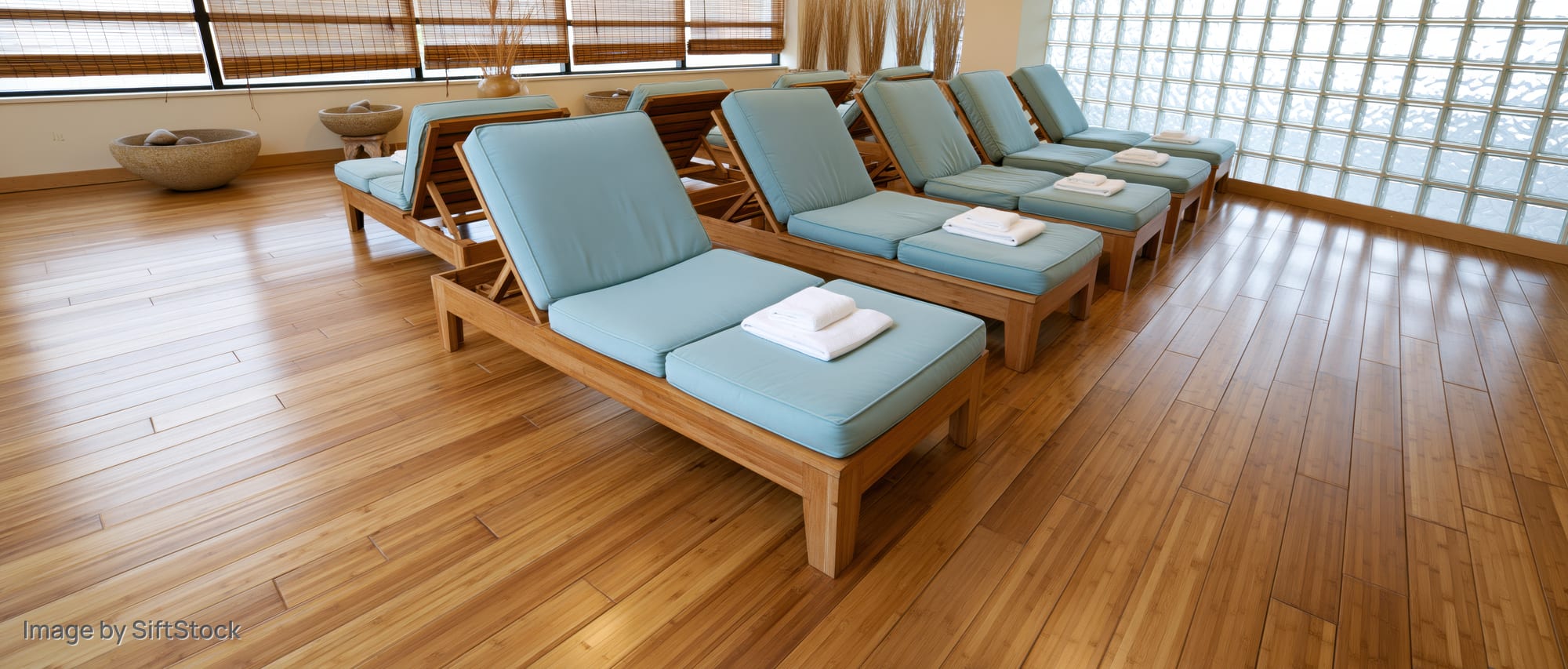 Row of wooden lounge chairs with blue cushions and white towels arranged in a spa relaxation room with hardwood floors and glass block windows