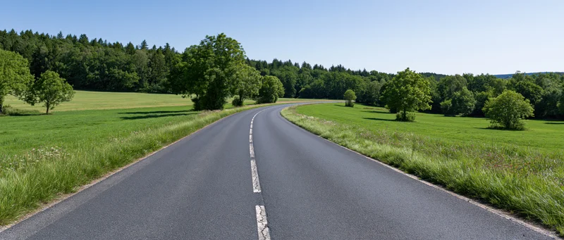 Winding asphalt road curving through green fields and trees under a clear blue sky