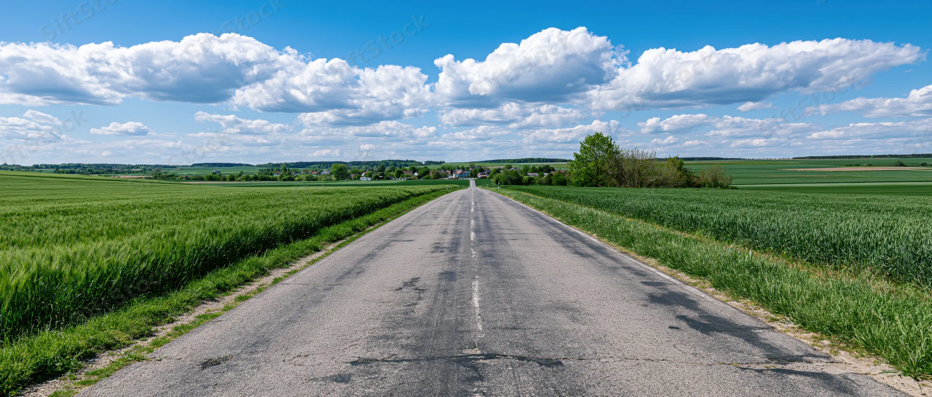 Straight asphalt country road stretching through green fields under a blue sky with white clouds