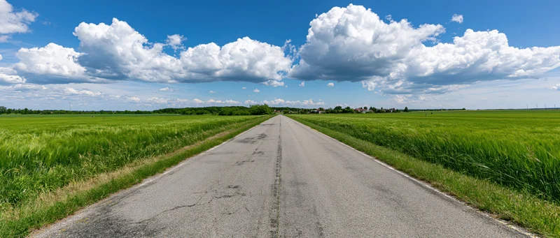 Straight asphalt country road stretching through green fields under blue sky with white clouds