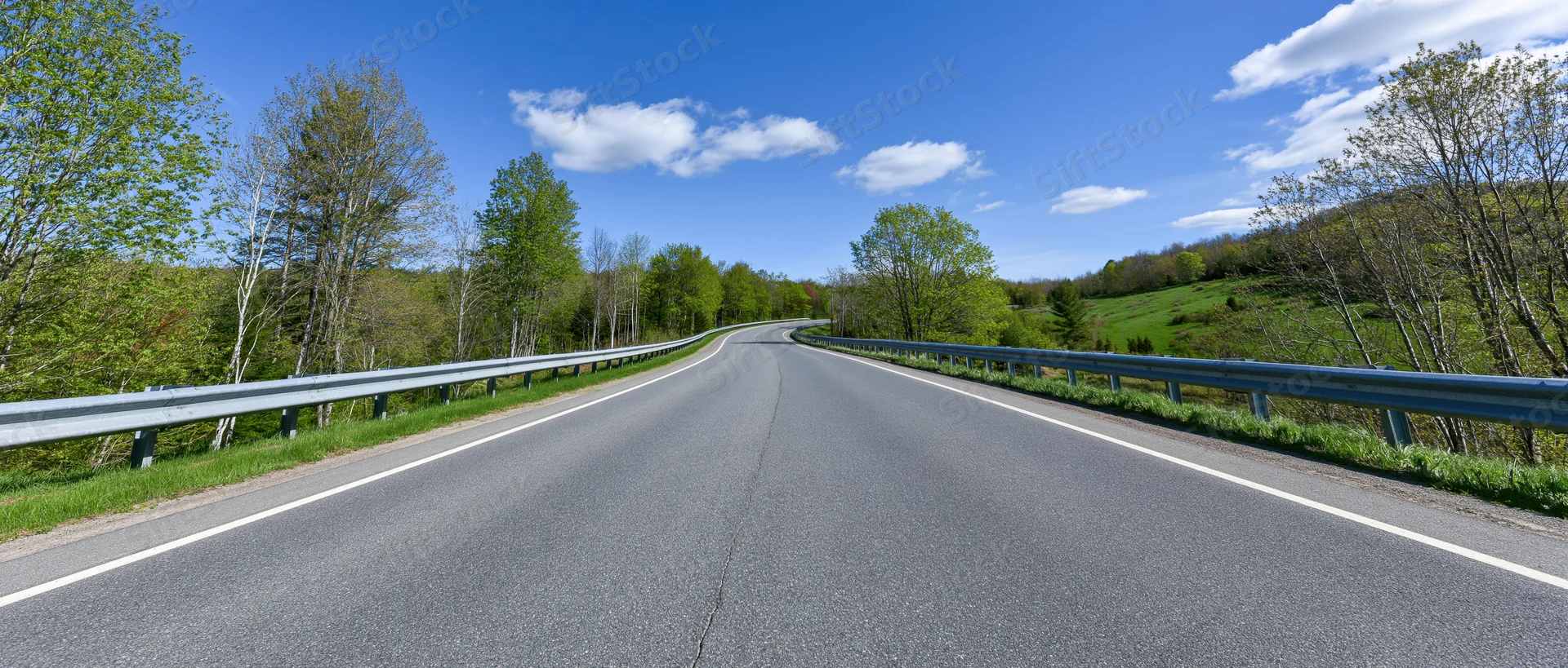 Empty two-lane asphalt highway curving gently to the left through green rural landscape under blue sky with clouds