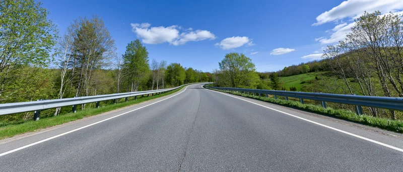 Empty two-lane asphalt highway curving gently to the left through green rural landscape under blue sky with clouds