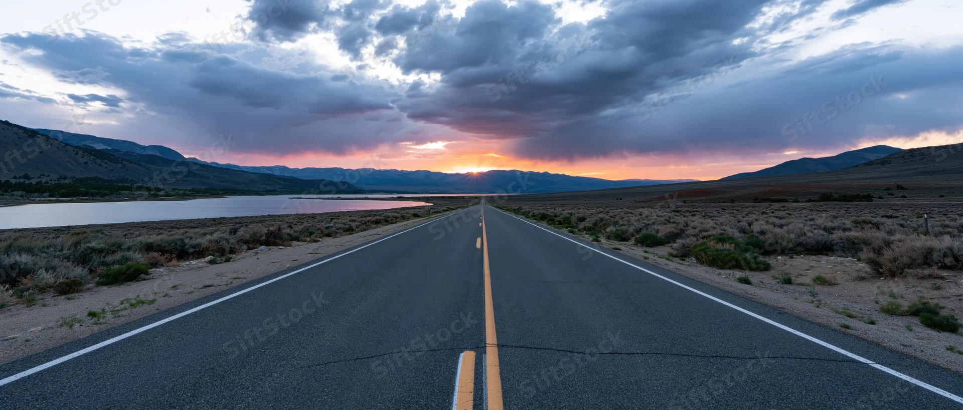 Straight asphalt highway stretching toward sunset over mountains and lake