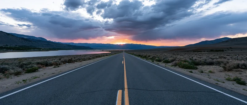 Straight asphalt highway stretching toward sunset over mountains and lake