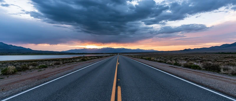 Straight asphalt highway stretching toward distant mountains at sunset over desert landscape