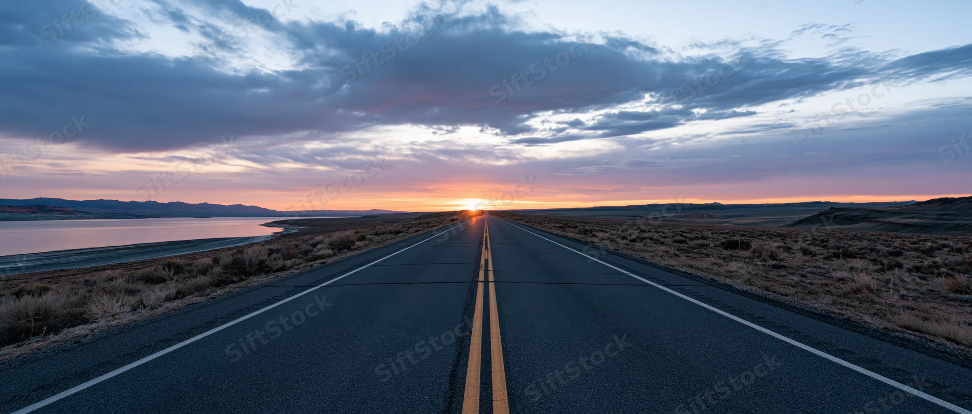 Straight two-lane asphalt highway stretching toward sunset over desert landscape with lake and mountains in distance