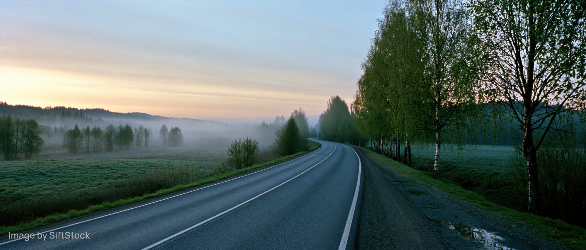 Curving two-lane asphalt road through misty rural landscape at dawn