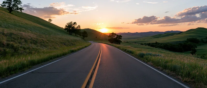Curving two-lane asphalt road through rolling green hills at sunset
