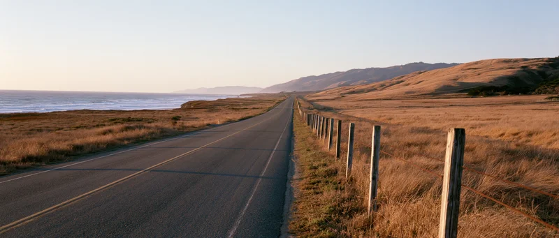 Two-lane highway stretching along the coast with dry grass fields and distant mountains under golden hour light