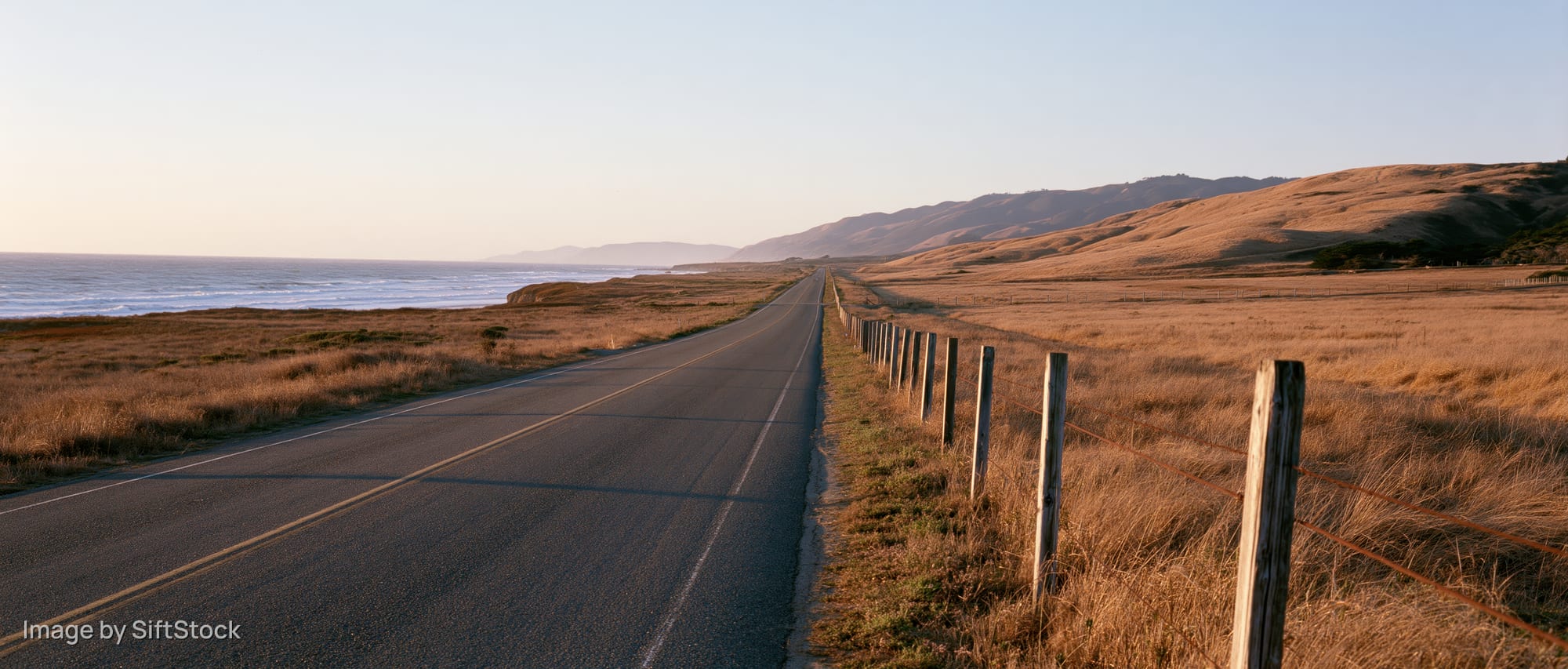 Two-lane highway stretching along the coast with dry grass fields and distant mountains under golden hour light