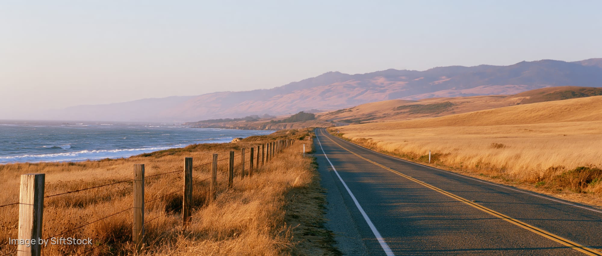 Two-lane highway stretching along the coast with ocean on one side and grassy hills on the other under golden hour light
