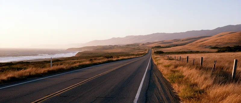 Two-lane highway stretching along the coast at sunset with golden grass fields and distant mountains