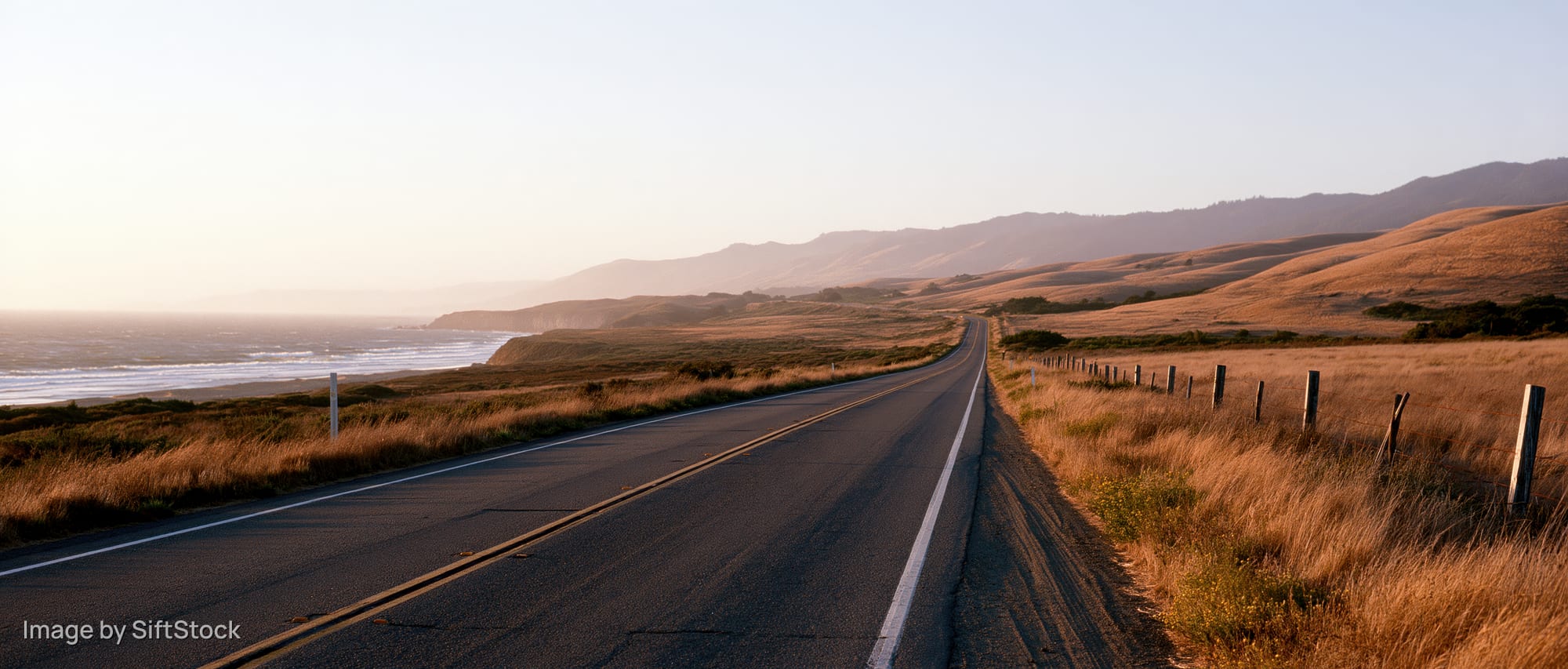 Two-lane highway stretching along the coast at sunset with golden grass fields and distant mountains