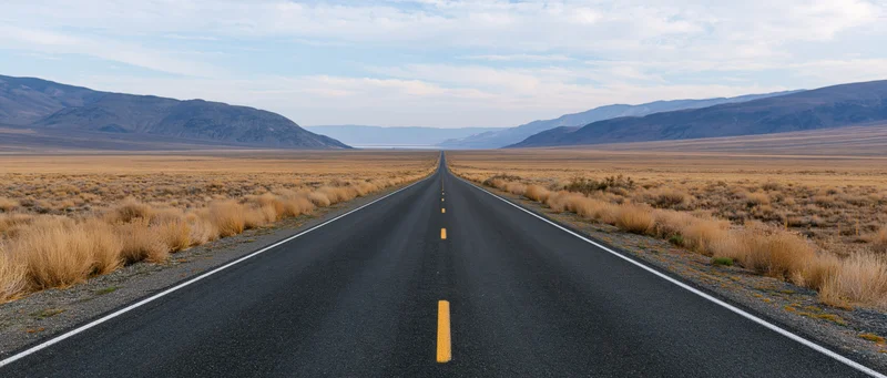 Straight asphalt highway through arid desert landscape with distant mountains under partly cloudy sky