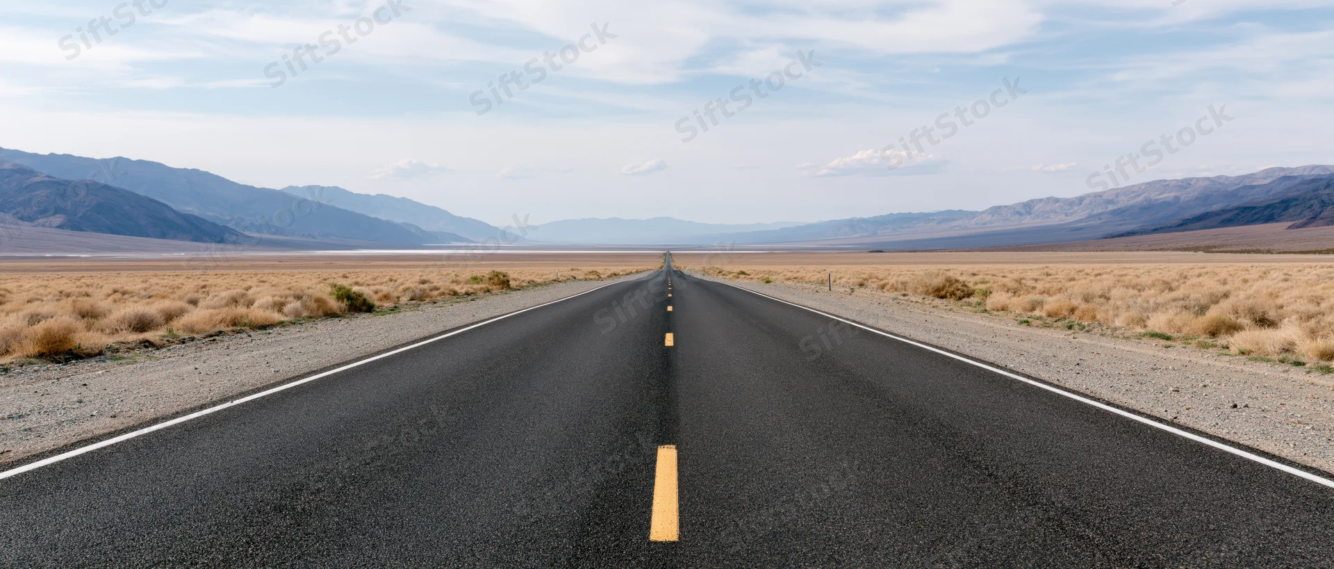 Straight asphalt highway stretching through arid desert landscape toward distant mountains under blue sky