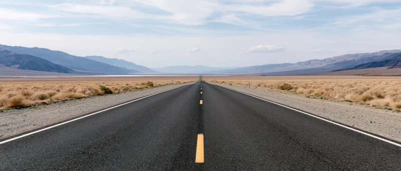 Straight asphalt highway stretching through arid desert landscape toward distant mountains under blue sky