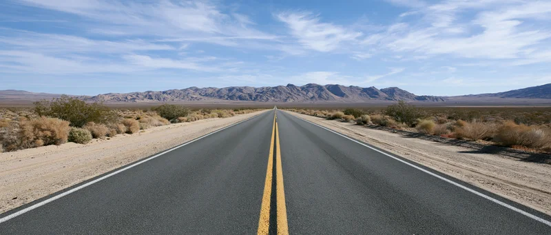 Straight two-lane highway stretching through desert landscape toward distant mountains under blue sky with clouds
