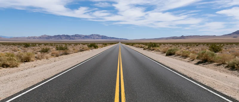 Straight asphalt highway stretching through arid desert landscape toward distant mountains under blue sky with clouds