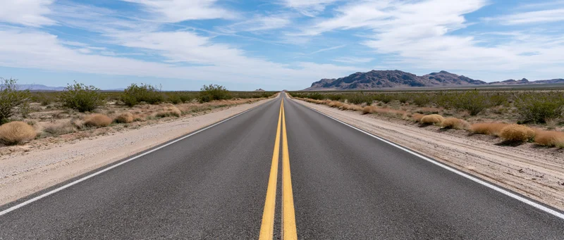 Straight asphalt highway stretching through arid desert landscape toward distant mountains under blue sky with clouds