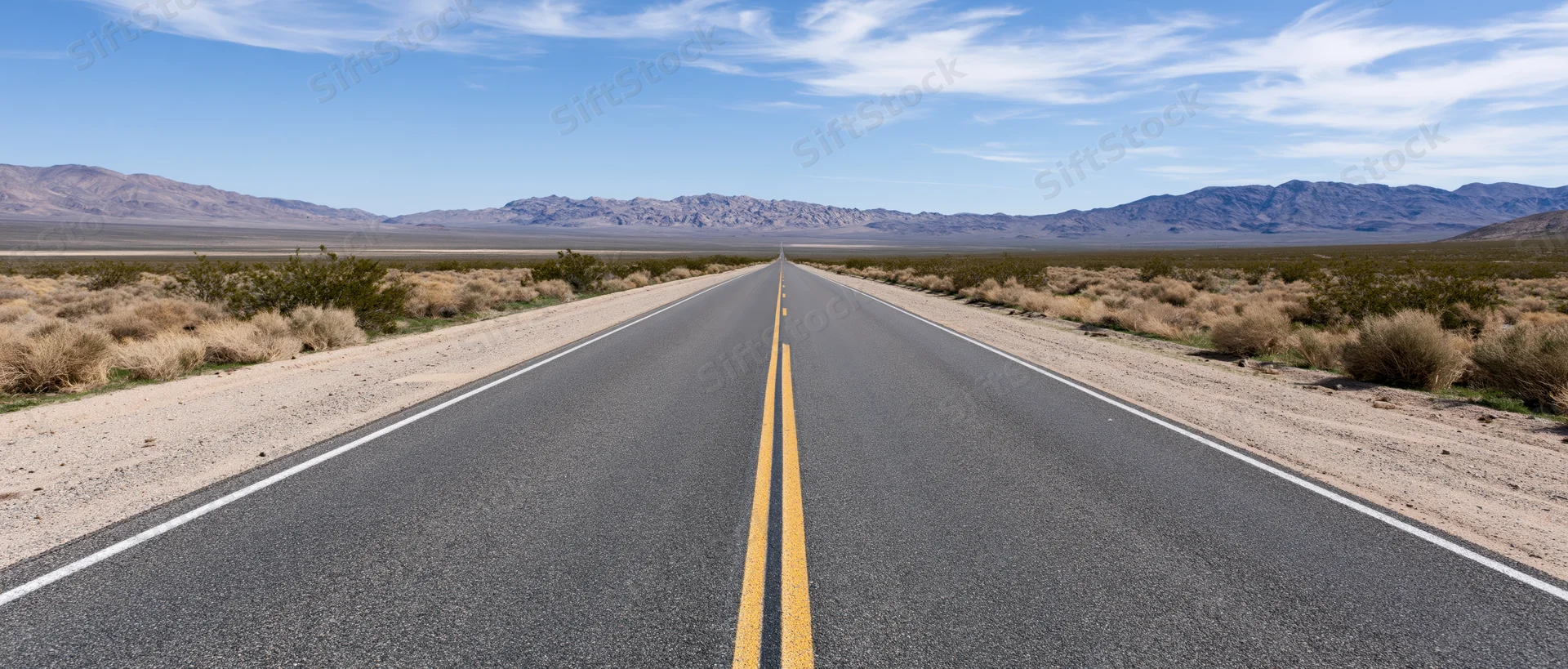 Straight two-lane highway stretching through desert landscape toward distant mountains under clear blue sky