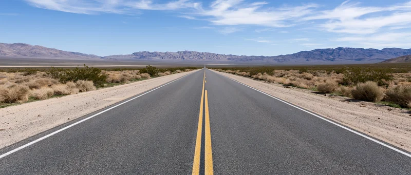 Straight two-lane highway stretching through desert landscape toward distant mountains under clear blue sky