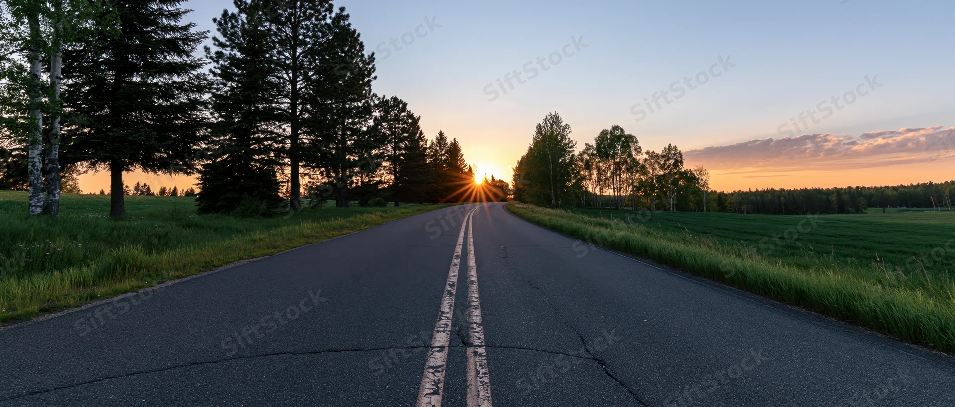 Two-lane asphalt road stretching toward sunset over rural landscape with trees and fields on either side