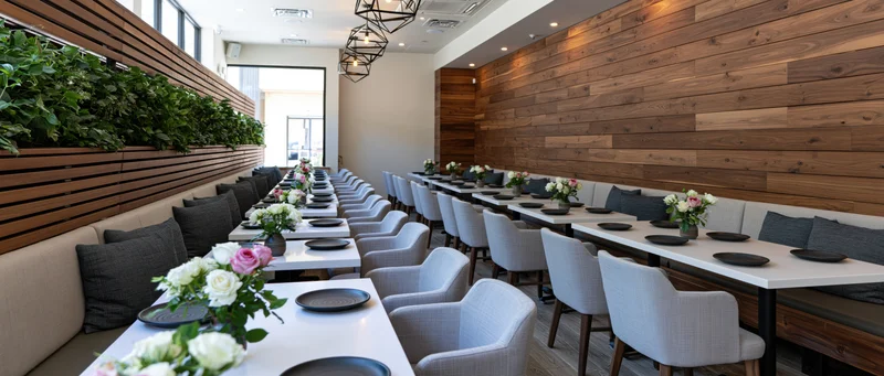 Long white tables set with black plates and floral arrangements in a modern restaurant dining room with wood walls and booth seating