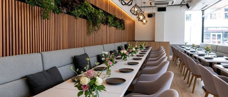 Long white tables set with black plates and floral centerpieces in a modern restaurant dining room with wooden walls and gray booths